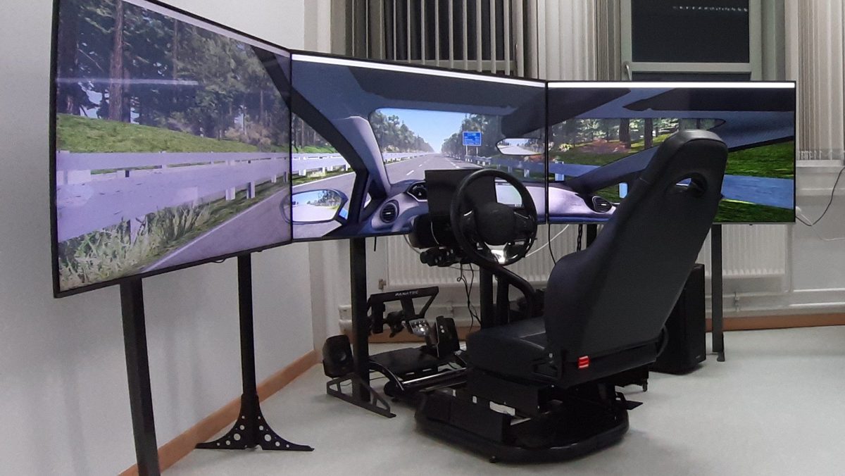 The driving simulator of the lab: a seat with steering wheel and pedals in front of three screens with a driving scene.